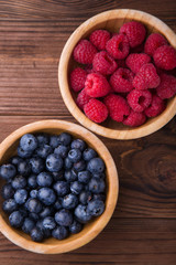 Two wooden bowls with raspberries and blueberries standing on brown wooden background. Summer fruits and berries. Healthy food
