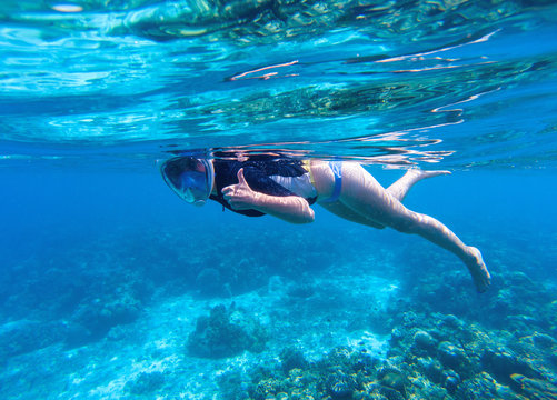 Woman snorkeling in blue water. Snorkel shows thumb in full face mask.