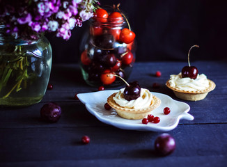 Mini tarts with whipped cream decorated with cherries, with a bouquet of flowers on a dark wooden background. Sweet cherries in a glass vase. Healthy deserts.