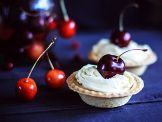 Mini tarts with whipped cream decorated with cherries, with a bouquet of flowers on a dark wooden background. Sweet cherries in a glass vase. Healthy deserts.