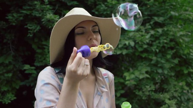 Slow motion of smiles young beautiful happy woman in hat blowing bubble in the park