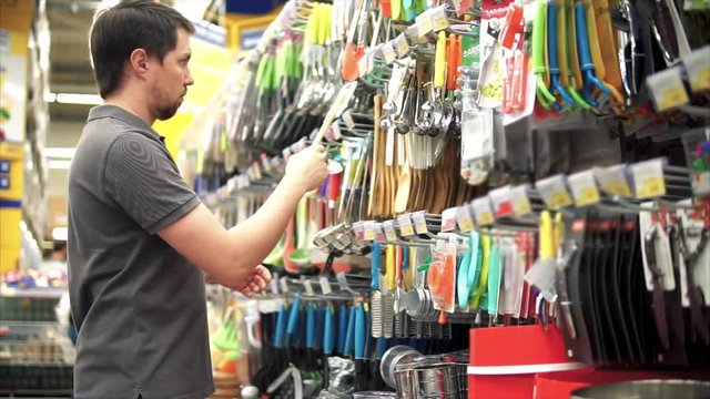 Male customer buying wooden spatula to his kitchen. He taking it from the shelf and holding it in hands. Man householder