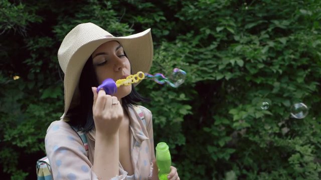 Slow motion of smiles young beautiful happy woman in hat blowing bubble in the park