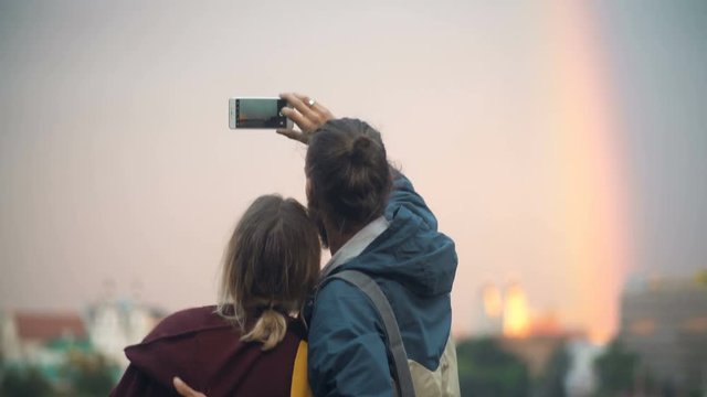 Back view of young beautiful couple taking pictures of city centre landscape, sunset and rainbow on smartphone.