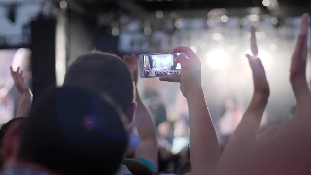 fans shoots video using mobile phone on concert, male relieves stage light with aid android at rock concert, people illuminated by colorful light during party clap their hands