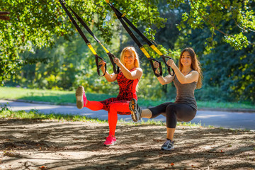 Two smiley beautiful girlfriends do crouches with special suspended device outdoor under the tree happy athletic blonde and brunette training in the park with fitness straps.
