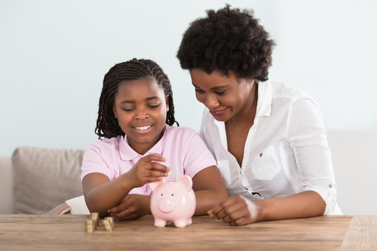 Girl With Mother Inserting Coins In Piggy Bank