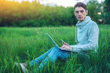 Naklejka na ściany i meble Young man sitting on a green meadow with laptop and using the phone on the background of blue cloudy sky at sunset