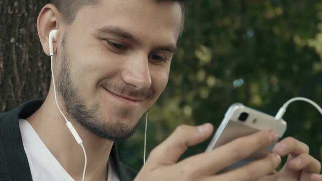 Smiling young man in the park . Browsing smartphone, listening to the music. People stock footage shot at summer season time. 