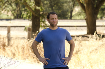 Man alone with hands on his hips in a natural outdoor setting, with trees and train tracks in the background. 