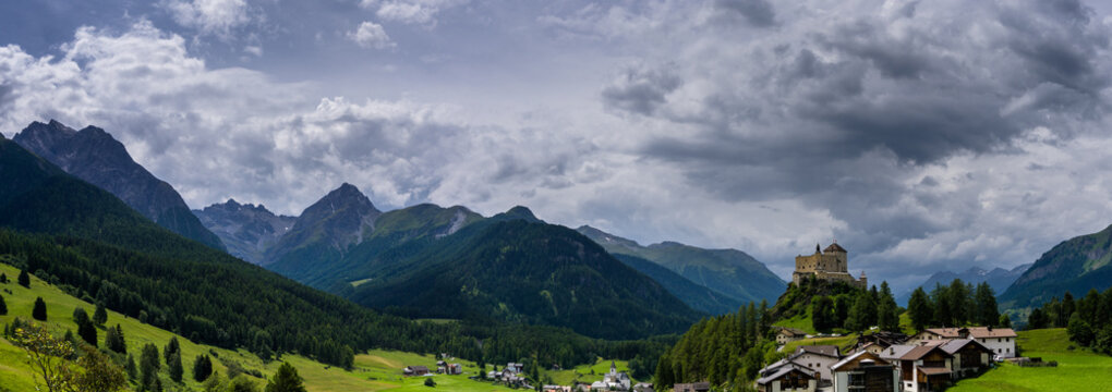 panorama view ofmountain peaks and a valley in the Swiss Alps with a small village and castle in the foreground underneath h a cloudy sky