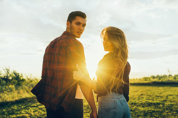 Rear view of awesome couple in a field sunset background. Handsome guy in plaid shirt hugs a girl with blond hair in plaid shirt look back. Lifestyle and travel concept.