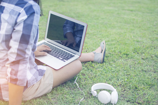 Trendy Hipster Asain Woman Wearing A Shirt Sitting On The Green Grass While Typing Thai Keyboard Of Laptop Put On The Leg With The Ear Plug For Relaxing In The Vacation