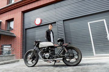 Handsome rider man in black leather biker jacket look to his classic style cafe racer motorcycle industrial gates as background. Bike custom made in vintage garage. Brutal fun urban lifestyle.