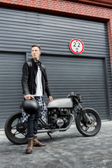 Handsome rider man in black leather biker jacket and boots near classic style cafe racer motorcycle industrial gates as background. Bike custom made in vintage garage. Brutal fun urban lifestyle.
