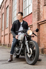Fototapeta na wymiar Handsome rider biker guy in leather jacket sit on classic style cafe racer motorcycle and look to the side. Bike custom made in vintage garage. Brutal fun urban lifestyle. Outdoor portrait.