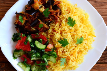 Simple vegetable risotto, stewed eggplants, salad with tomatoes and cucumbers on a white plate and a wooden table. Vegetarian dish idea. Closeup