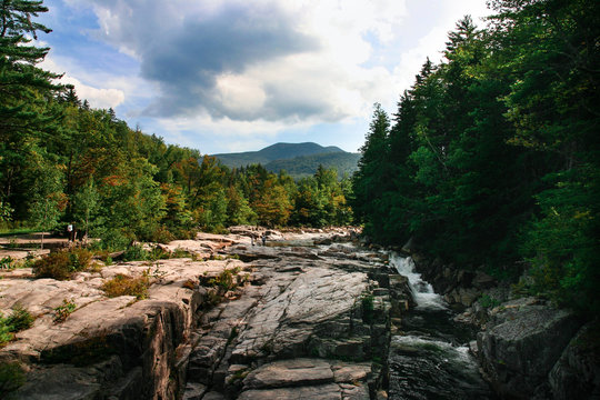 River in New Hampshire flanked by rocks and trees with blue sky and some clouds in the background