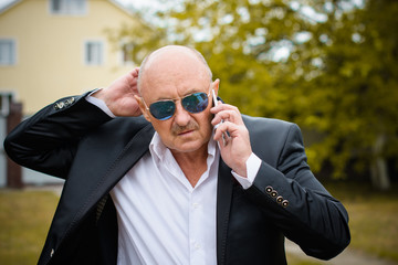 Portrait stylish mature men in white shirt and jacket and blue sunglasses using smartphone. Serving internet via his gadget. Old man using social network technology. Senior man talking on the phone