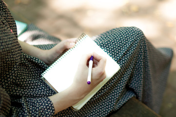 Legs of a  woman in long dress, on a bench in the park. Girl Write notes to notepad in a park outdoors Close-up. Point of view.