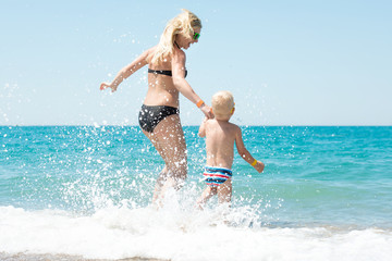 Naklejka premium Beautiful young mother and small son holding hands running on the waves on the beach.Fun,family,friendly summer vacation.