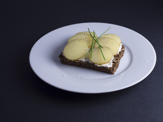 Danish open faced sandwich (smoerrebroed) with smoked cheese, sliced new potatoes and chives on dark rye bread. Placed on white plate on a black background.