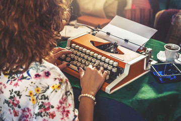 Young woman,retro look, sitting at a typewriter, portrait. Added noise. Photographed from behind, hands on a typewriter