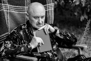 Portrait of old elegant man with gray hair reading a book in plaid armchair outside in park and thinking about his life