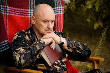 Portrait of old elegant man with gray hair reading a book in plaid armchair outside in park and thinking about his life
