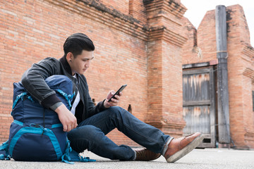 young traveler, asian man wearing black jacket and blue jeans sitting near old orange brick wall with mobile smart phone and backpack