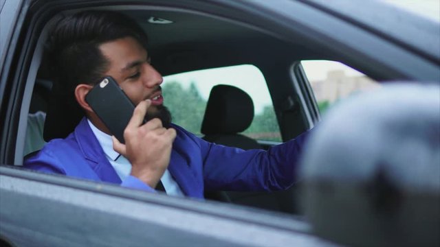 Modern arab man in formal wear sitting in right-hand drive car on the driver's seat and talking on cell phone
