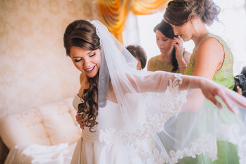 Stylish beautiful bridesmaids helping gorgeous brunette bride in white dress get ready for wedding, morning preparations, woman putting on dress