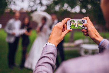 Man taking photo on phone of stylish wedding bride and groom posing. photo booth. wedding couple making photos with friends on phone camera. hand holding smartphone