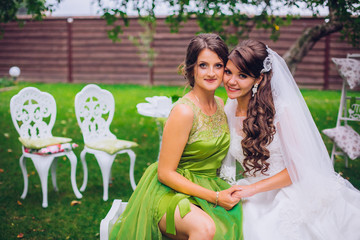 Bride and bridesmaid embracing in the park on the wedding day. Bridesmaid in green color dress.