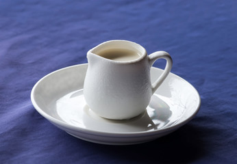 White ceramic creamer on a saucer with cold cream on a blue background.