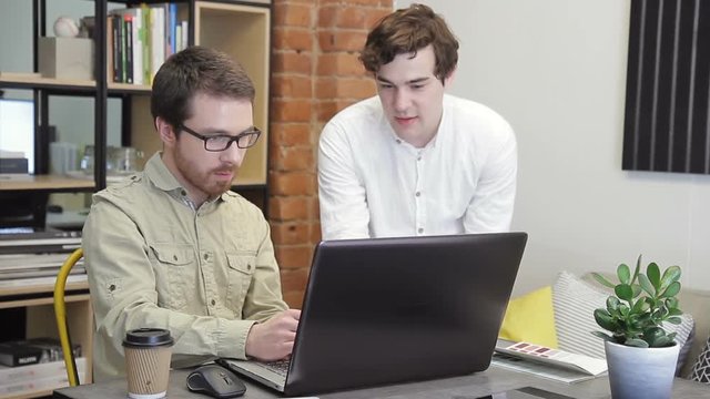 In the office, two young men work in front of the computer. The guy with the beard and with glasses carefully looks at the monitor. The second one sits next to it and points to the screen with a pen