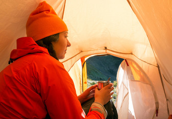 Cute young woman sits in a tent in an orange jacket and hats, holding a cup in her hands.