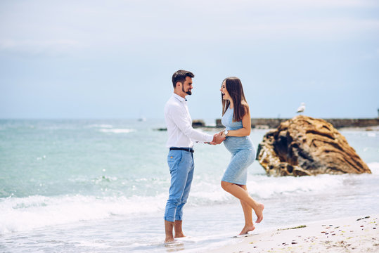 A beautiful smiling pregnant woman in a blue dress and her courageous and handsome husband in a shirt and blue trousers leaned to each other against the background of the rocks and the sea.