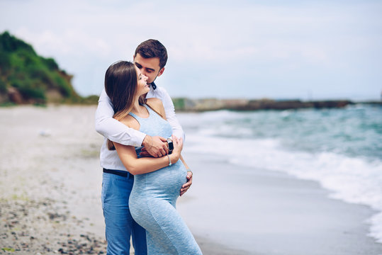 Courageous and handsome man in a white shirt and blue trousers  hugs and kissing on a cheek her beautiful pregnant wife in a blue dress against the background of the sea.