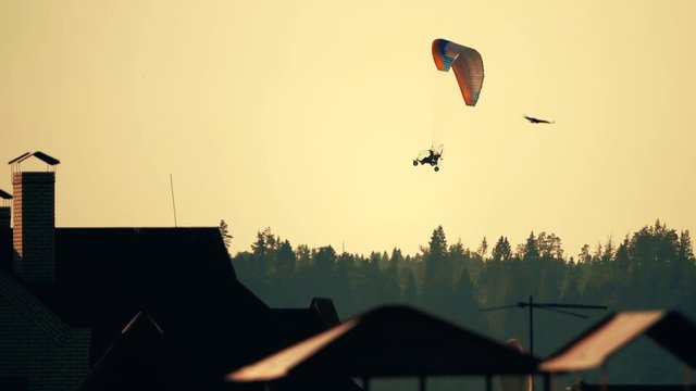 Unknown powered paraglider flying above the forest and town rooftops at sunset. Slow motion shot