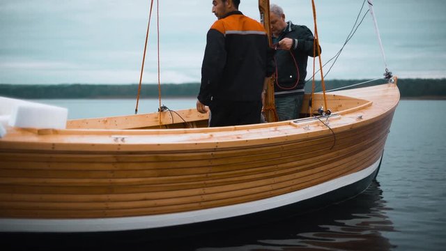 Mature Master and his asian assistant working on wooden sailboat on river or lake. Smooth movement footage. They try to install mast. Close-up shooting teal and orange. 4k ultrahd