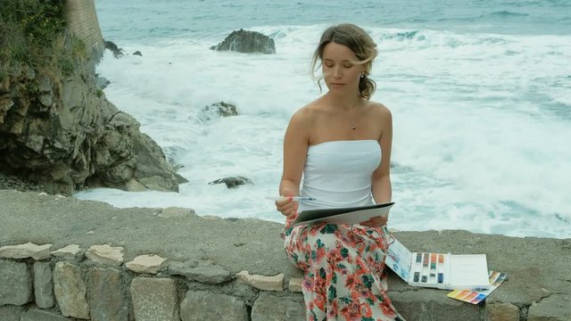 Young woman sits on seashore and paints with watercolors outside. Attractive blond female with curly dyed hair, sits on stones coast in white T-shirt, colored skirt and holds paper in her hands