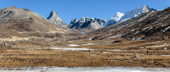 Mountains witn snow and below with tourists on the ground with brown grass, snow and frozen pond in winter at Zero Point at Lachung. North Sikkim, India.