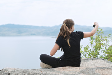 Naklejka na ściany i meble The girl makes selfie/The girl sits on the shore of the lake and makes a selfie, the view from the back