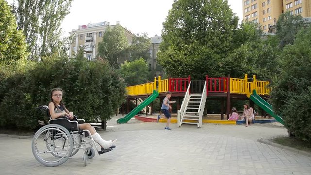 A girl with a broken leg sits in a wheelchair in front of the playground