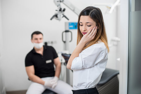 Beautiful woman suffering from terrible teeth pain, touching cheek with hand at dental clinic. Female feeling toothache. On the background dentist sitting on the chair. Dental care and health concept