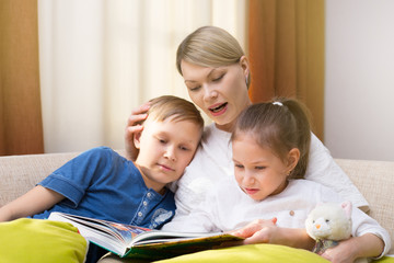 Obraz premium Beautiful mother is reading a book to her young children. Sister and brother is listening to a story. Happy loving family.