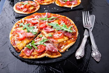 Homemade pizza with prosciutto, tomato, arugula on black slate board. Close up.