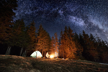 Male tourist enjoying in his camp near the forest at night. Man sitting near campfire and tent under beautiful night sky full of stars and milky way. Astrophotography