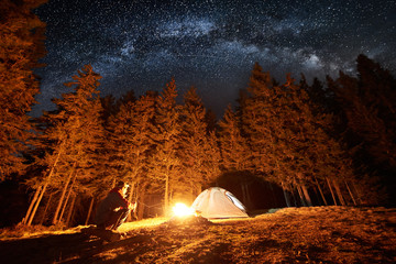 Male tourist enjoying in his camping near the forest at night. Man sitting near campfire and tent under beautiful night sky full of stars and milky way. Long exposure